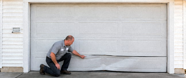 Garage door technician inspecting a bent lower panel on a white residential garage door in Sacramento, CA.